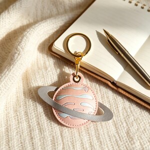 May include: A pink planet-shaped keychain with a gray ring, gold-colored hardware, and a matching clasp. The keychain is made of leather and has light blue and white accents. The keychain is lying on a white knitted surface.