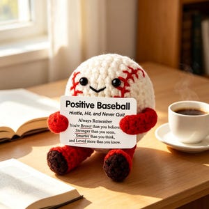 Puede incluir: Un peluche de béisbol de ganchillo redondo y color crema con costuras rojas, ojos de cuentas negras y una pequeña sonrisa. Tiene brazos y piernas de ganchillo rojos y sostiene un cartel blanco que dice: Positive Baseball, Hustle, Hit, and Never Quit.