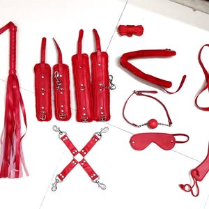 May include: A collection of red accessories including a multi-strand whip, four padded wrist and ankle cuffs with metal buckles, a cross-shaped strap connector, a ball gag, a blindfold, a heart-shaped paddle, a leash, and a coiled rope.
