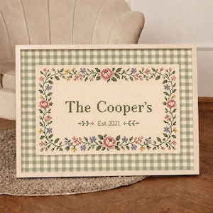 May include: A rectangular framed cross-stitch sampler featuring a light green and white gingham border. The centre displays the text The Cooper's and Est. 2021, surrounded by a floral wreath of pink roses and small blue and yellow flowers.