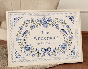 May include: A rectangular framed cross-stitch sampler on a light fabric background featuring a blue floral wreath, two blue birds, a central urn, and a blue bow. The text reads The Andersons and Est. 2026 in blue thread within the floral border.