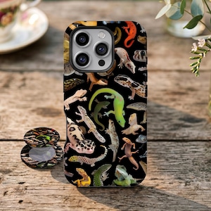 Könnte beinhalten: Eine schwarze Smartphone-Hülle und ein passender runder Handyhalter mit einem sich wiederholenden Muster aus verschiedenen bunten Eidechsen, darunter Leopardengeckos, grüne Geckos und orangefarbene Salamander, die über die gesamte Oberfläche verteilt sind.