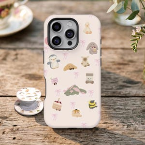 Peut inclure: Une coque de smartphone rose pâle et un support de téléphone circulaire assorti présentant un motif répétitif de petites illustrations mignonnes, dont un pingouin, un lapin, une abeille, un croissant, un gâteau et des nœuds roses.