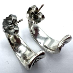 May include: A pair of silver-toned, curved stud earrings. The earrings have a smooth, polished finish and a post and butterfly backing. The design is a simple, elegant crescent shape, suitable for everyday wear. The earrings are displayed on a white surface.