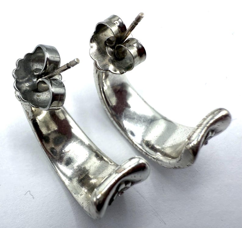 May include: A pair of silver-toned, curved stud earrings. The earrings have a smooth, polished finish and a post and butterfly backing. The design is a simple, elegant crescent shape, suitable for everyday wear. The earrings are displayed on a white surface.