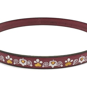 May include: A burgundy bangle bracelet with a repeating floral pattern in white and gold. The bracelet is a simple band, and the floral design is evenly spaced around the circumference.