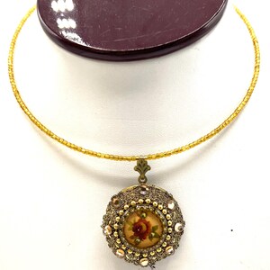May include: A gold-toned necklace with a pendant featuring a miniature rose painting. The pendant is round, framed with gold beads and crystals. The necklace has a delicate beaded chain.