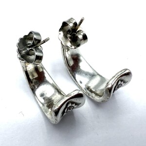 May include: A pair of silver-toned hoop earrings. Each earring has a curved, crescent shape with a small floral design at the bottom. The earrings have a post and butterfly backing for secure wear. The earrings are displayed on a white background.