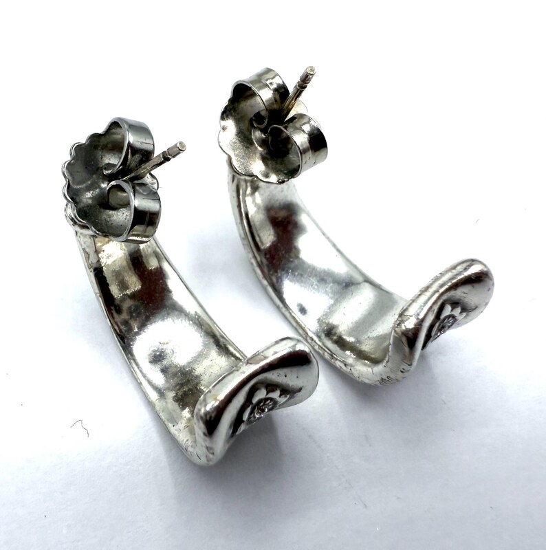 May include: A pair of silver-toned hoop earrings. Each earring has a curved, crescent shape with a small floral design at the bottom. The earrings have a post and butterfly backing for secure wear. The earrings are displayed on a white background.