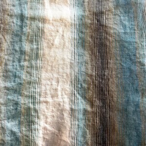 May include: Close-up of a textured fabric with vertical stripes in shades of blue, beige, and brown. The fabric has a slightly wrinkled appearance, suggesting it may be linen or a similar material. The stripes vary in width and colour intensity.