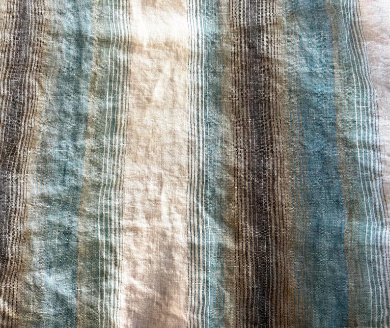 May include: Close-up of a textured fabric with vertical stripes in shades of blue, beige, and brown. The fabric has a slightly wrinkled appearance, suggesting it may be linen or a similar material. The stripes vary in width and colour intensity.