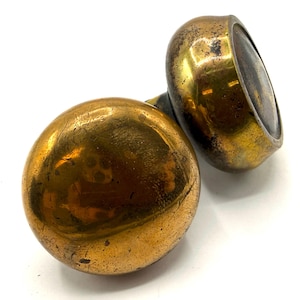 May include: Two antique brass door knobs. One knob is spherical, the other is a flattened oval shape. Both knobs have a tarnished, aged appearance with visible wear and tear, suggesting they are vintage.