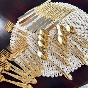 May include: A set of gold-toned flatware including knives, forks, and spoons with ornate, embossed handles. The cutlery is arranged on a round, cream-colored crocheted placemat, showcasing a polished metallic finish and detailed decorative patterns.