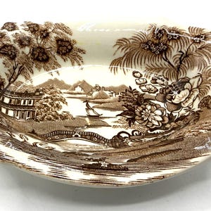 May include: Oval-shaped ceramic dish with a brown transferware landscape design. The dish features a detailed scene of a bridge, buildings, trees, and a boat on a body of water. The rim has two small handles. The color palette is primarily brown and cream.