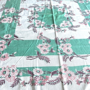 May include: A rectangular tablecloth featuring a white base with wide, solid mint green bands arranged in a grid pattern. The fabric is decorated with repeating floral motifs of pink daisy-like flowers and dark purple foliage.