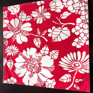 May include: Square red and white floral placemat. The placemat features a repeating pattern of large white flowers and leaves on a red background. The edges are neatly stitched.