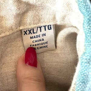 May include: A close-up of a clothing label showing "XXL/TTG MADE IN CHINA FABRIQUE EN CHINE". The label is attached to a beige fabric, with a section of light blue fabric on the right.
