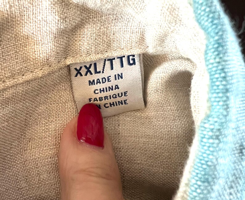 May include: A close-up of a clothing label showing "XXL/TTG MADE IN CHINA FABRIQUE EN CHINE". The label is attached to a beige fabric, with a section of light blue fabric on the right.