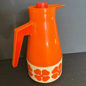 May include: An orange vintage thermos with a red lid and handle. The base features a cream-colored band with orange flower designs. The thermos is designed with a spout for easy pouring.