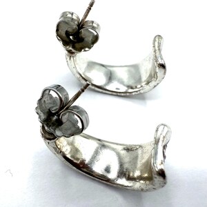 May include: A pair of silver-toned hoop earrings. Each earring has a curved, crescent shape with a post and butterfly backing. The earrings are displayed against a white background.