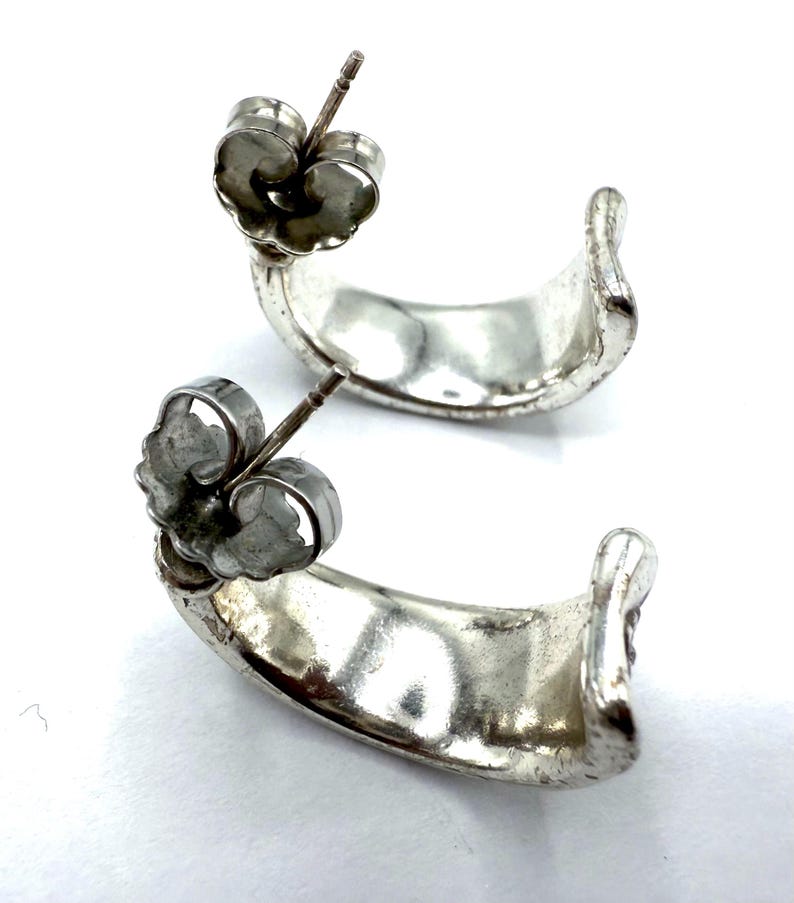 May include: A pair of silver-toned hoop earrings. Each earring has a curved, crescent shape with a post and butterfly backing. The earrings are displayed against a white background.