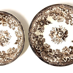 May include: Two ceramic plates with a floral pattern in brown and cream. The plates have a scalloped edge and a detailed floral design in the center. The plates are likely for serving food.