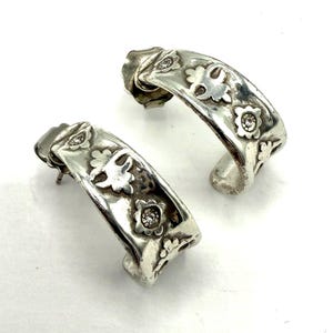 May include: A pair of silver-toned hoop earrings. Each earring features a floral design with small, clear gemstones. The earrings have a curved shape and a post closure. The design includes small flowers and cut-out details.
