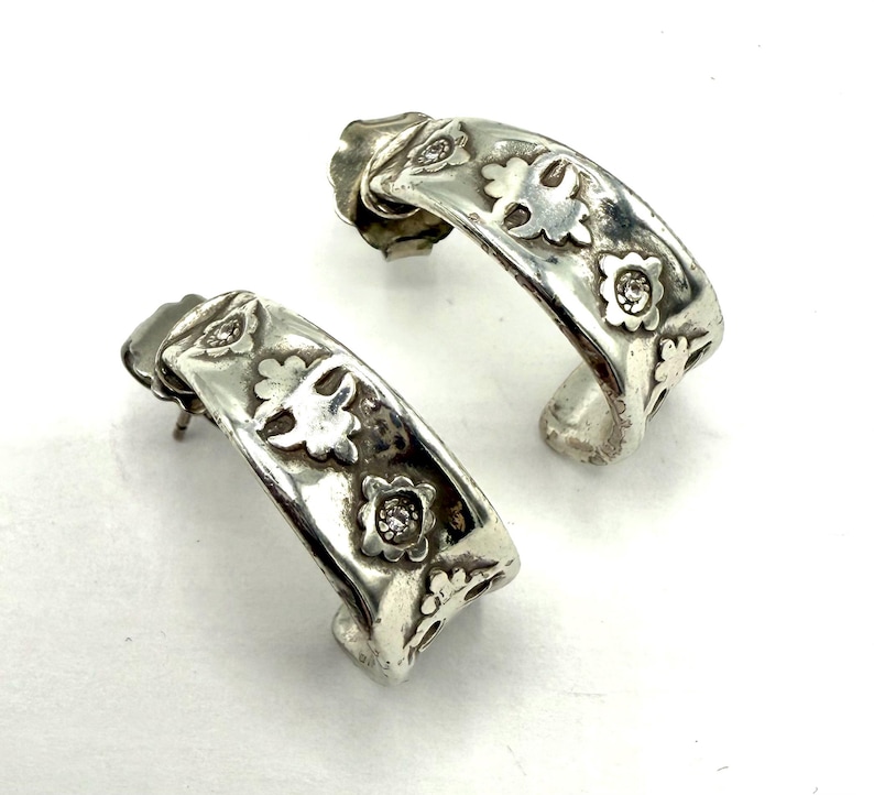 May include: A pair of silver-toned hoop earrings. Each earring features a floral design with small, clear gemstones. The earrings have a curved shape and a post closure. The design includes small flowers and cut-out details.