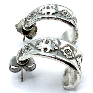 May include: A pair of silver hoop earrings. Each earring features a curved design with floral and leaf patterns, and small, clear gemstones. The earrings have a post and butterfly backing.