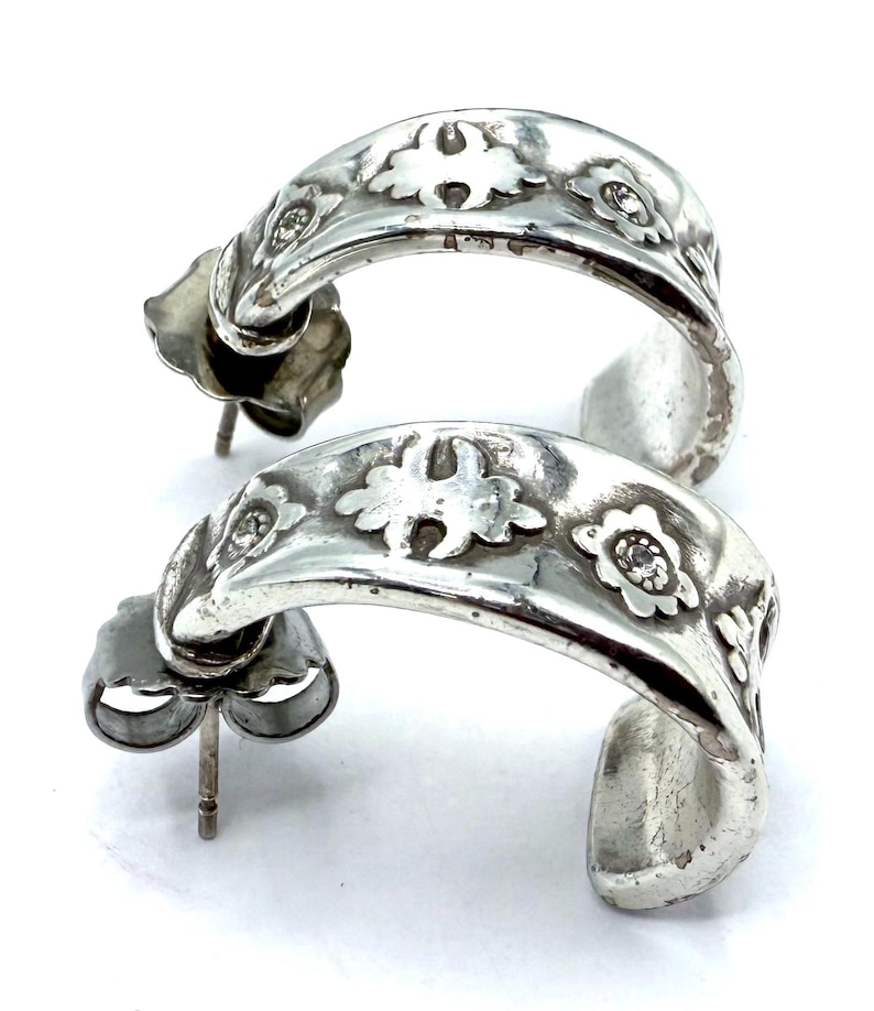 May include: A pair of silver hoop earrings. Each earring features a curved design with floral and leaf patterns, and small, clear gemstones. The earrings have a post and butterfly backing.