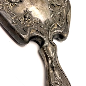 May include: Antique silver-toned hand mirror with floral and leaf detailing. The mirror's frame and handle feature an ornate, Art Nouveau-style design with raised relief flowers and flowing lines. The handle is curved and tapers towards the base.