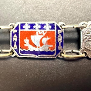 May include: A silver-toned metal charm bracelet featuring three rectangular links. The left link depicts the Arc de Triomphe, the center link displays the blue, red, and white enamel coat of arms of Paris with a ship, and the right link shows the Eiffel Tower.
