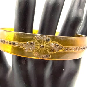 May include: A vintage, translucent amber-colored bangle bracelet. The bracelet features a floral design with clear and smoky-colored rhinestones. Additional rhinestones are set along the top and bottom edges of the bracelet.