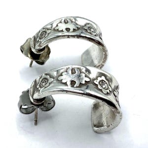 May include: A pair of silver-toned hoop earrings. Each earring features a curved design with embossed floral patterns and small, round embellishments. The earrings have post-and-clutch closures.