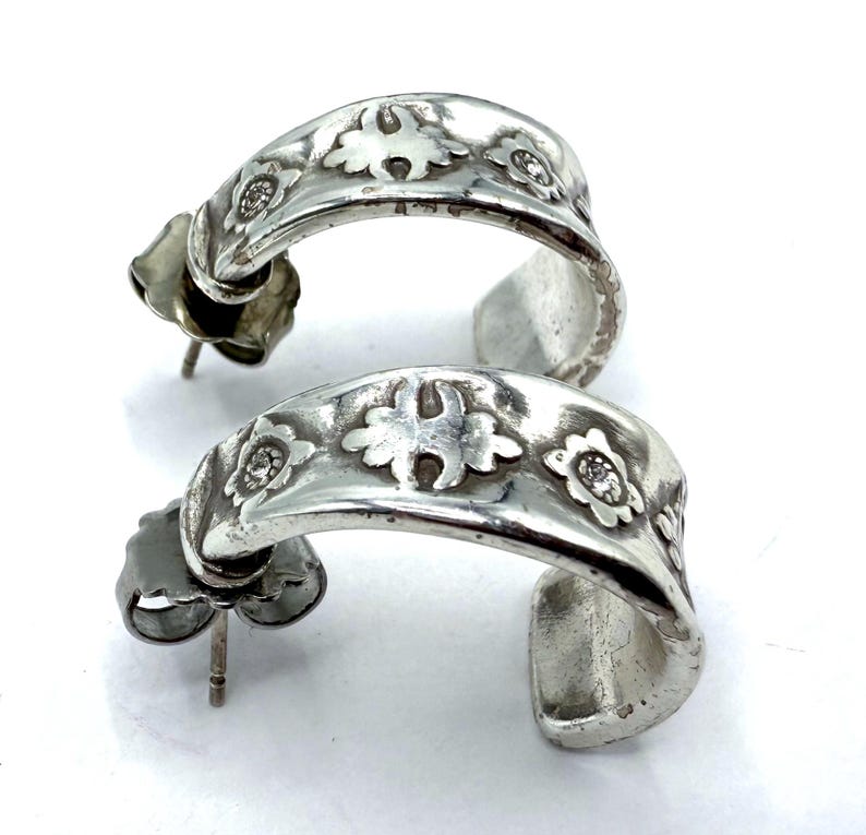 May include: A pair of silver-toned hoop earrings. Each earring features a curved design with embossed floral patterns and small, round embellishments. The earrings have post-and-clutch closures.
