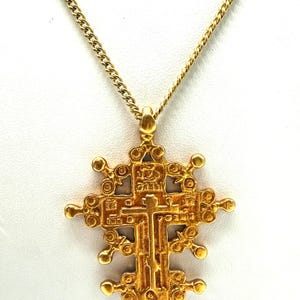 May include: Gold-toned pendant necklace featuring a detailed cross design. The cross has an ornate, textured surface with intricate patterns and small circular accents. The necklace chain is a delicate curb link style.