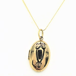 May include: An oval-shaped gold locket pendant suspended from a delicate gold chain. The locket features a brushed gold finish with a central black engraved shield-like motif and decorative floral accents at the top, secured by a small jump ring.