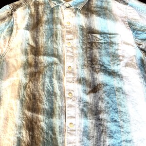 May include: A striped button-down shirt in blue, white, and brown. The shirt features a chest pocket and a collar. The brand name "Tommy Bahama" is visible on a tag near the collar.