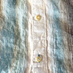 May include: Close-up of a striped shirt in shades of blue, beige, and grey. The fabric has a textured appearance with vertical stripes. The shirt features beige buttons down the centre.