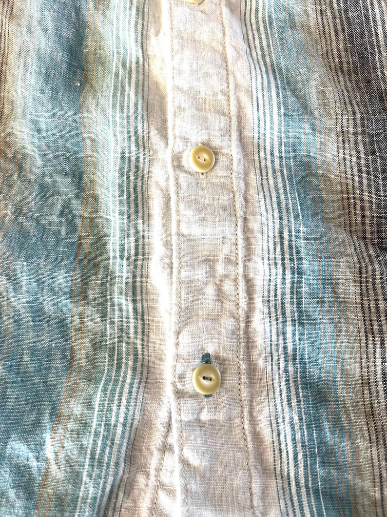 May include: Close-up of a striped shirt in shades of blue, beige, and grey. The fabric has a textured appearance with vertical stripes. The shirt features beige buttons down the centre.