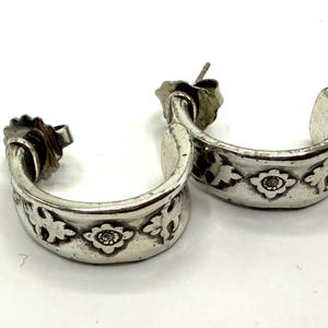 May include: A pair of silver-toned hoop earrings. Each earring features a floral design with a central, small, round embellishment. The earrings are on a white surface.