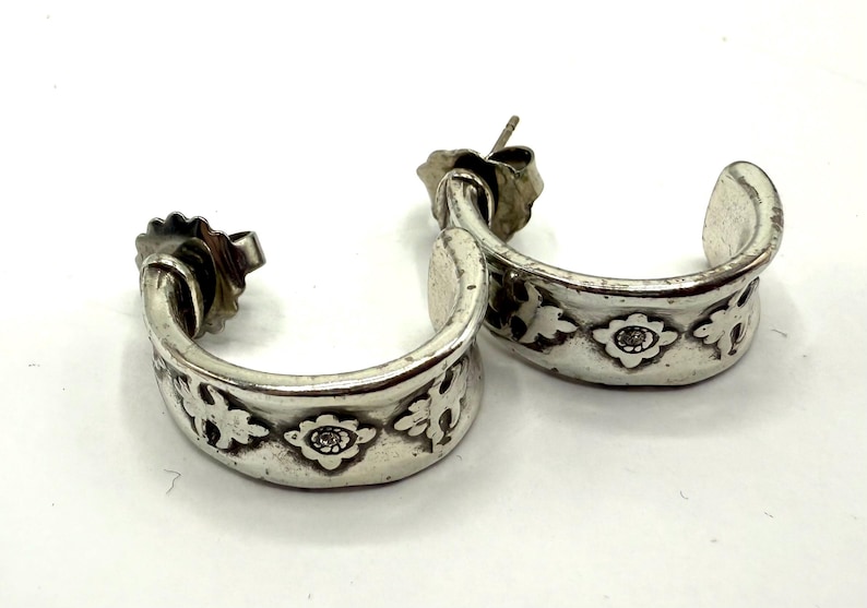 May include: A pair of silver-toned hoop earrings. Each earring features a floral design with a central, small, round embellishment. The earrings are on a white surface.