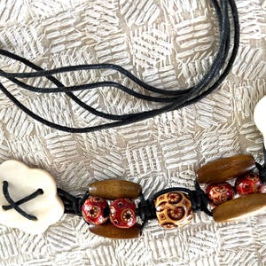 May include: A necklace featuring black cord strands accented with white flower-shaped components, brown elongated wooden beads, and smaller round beads with red and brown geometric patterns. The components are strung together with black cord.