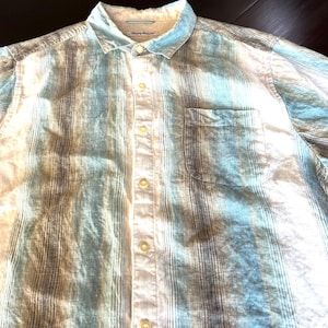 May include: A short-sleeved button-down shirt with a striped pattern in shades of blue, white, and grey. The shirt has a chest pocket and a classic collar. The fabric appears to be lightweight.