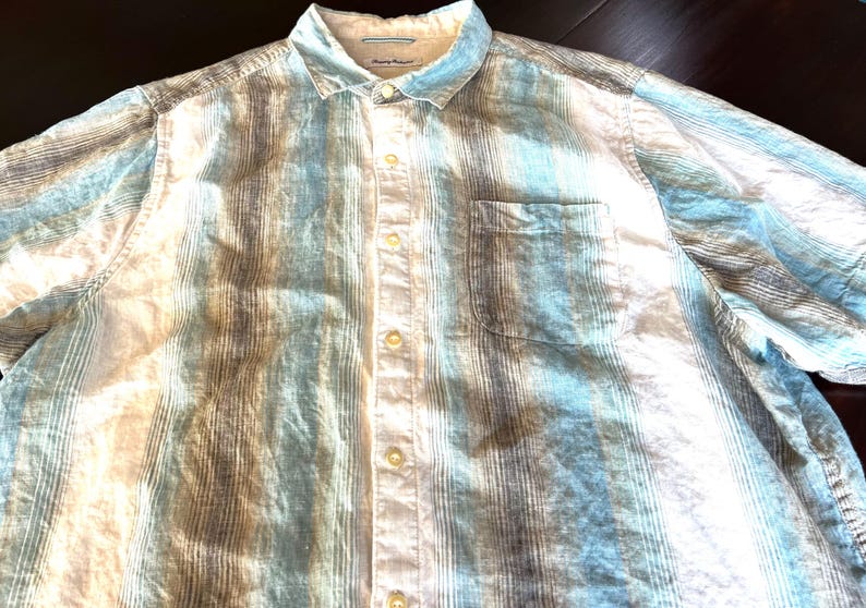 May include: A short-sleeved button-down shirt with a striped pattern in shades of blue, white, and grey. The shirt has a chest pocket and a classic collar. The fabric appears to be lightweight.