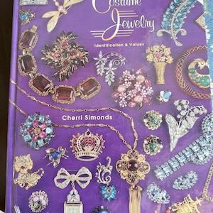 May include: A book titled "Collectible Costume Jewelry: Identification & Values" by Cherri Simonds. The cover is purple and features various pieces of vintage jewelry, including brooches, necklaces, and earrings. The book's cover also has the text "Values Updated 2003".