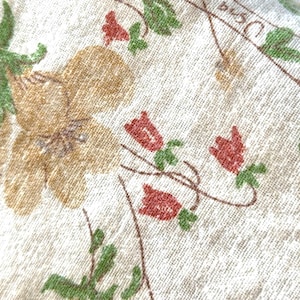 May include: Close-up of a cream-colored fabric with a floral pattern. The design features large tan flowers, small red buds, and green leaves. The fabric has a textured appearance.