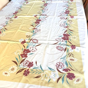 May include: A rectangular tablecloth featuring a white center and a wide mustard yellow border. The design includes a floral garland pattern with maroon, white, and teal flowers and green leaves arranged in a continuous rectangular frame.