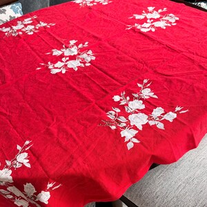May include: A rectangular red tablecloth featuring a repeating pattern of white floral clusters. The fabric displays a series of white rose-like blossoms and leaves distributed across the surface, creating a consistent decorative design.