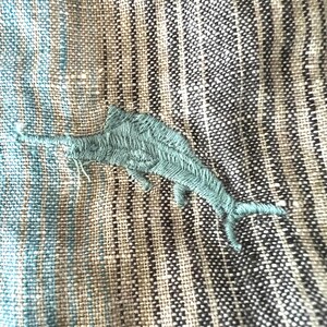 May include: Close-up of textured fabric featuring a teal embroidered marlin. The fabric has a striped pattern in shades of blue, beige, and black. The marlin is detailed with visible stitching, adding texture to the design.